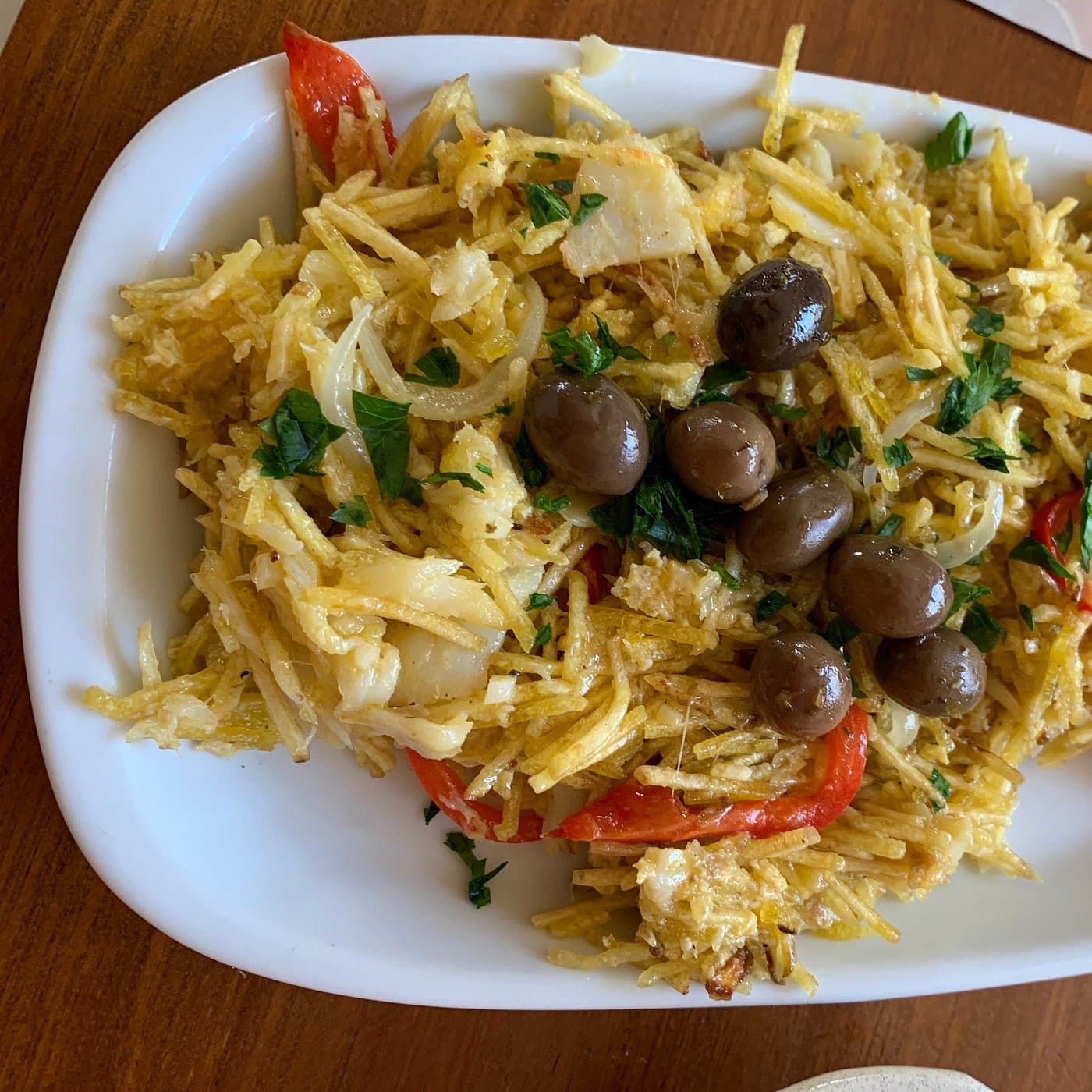 Bacalhau à Brás numa travessa branca: batata palha, lascas de bacalhau, cebola, salsa picada, tiras de pimento vermelho e azeitonas pretas por cima.