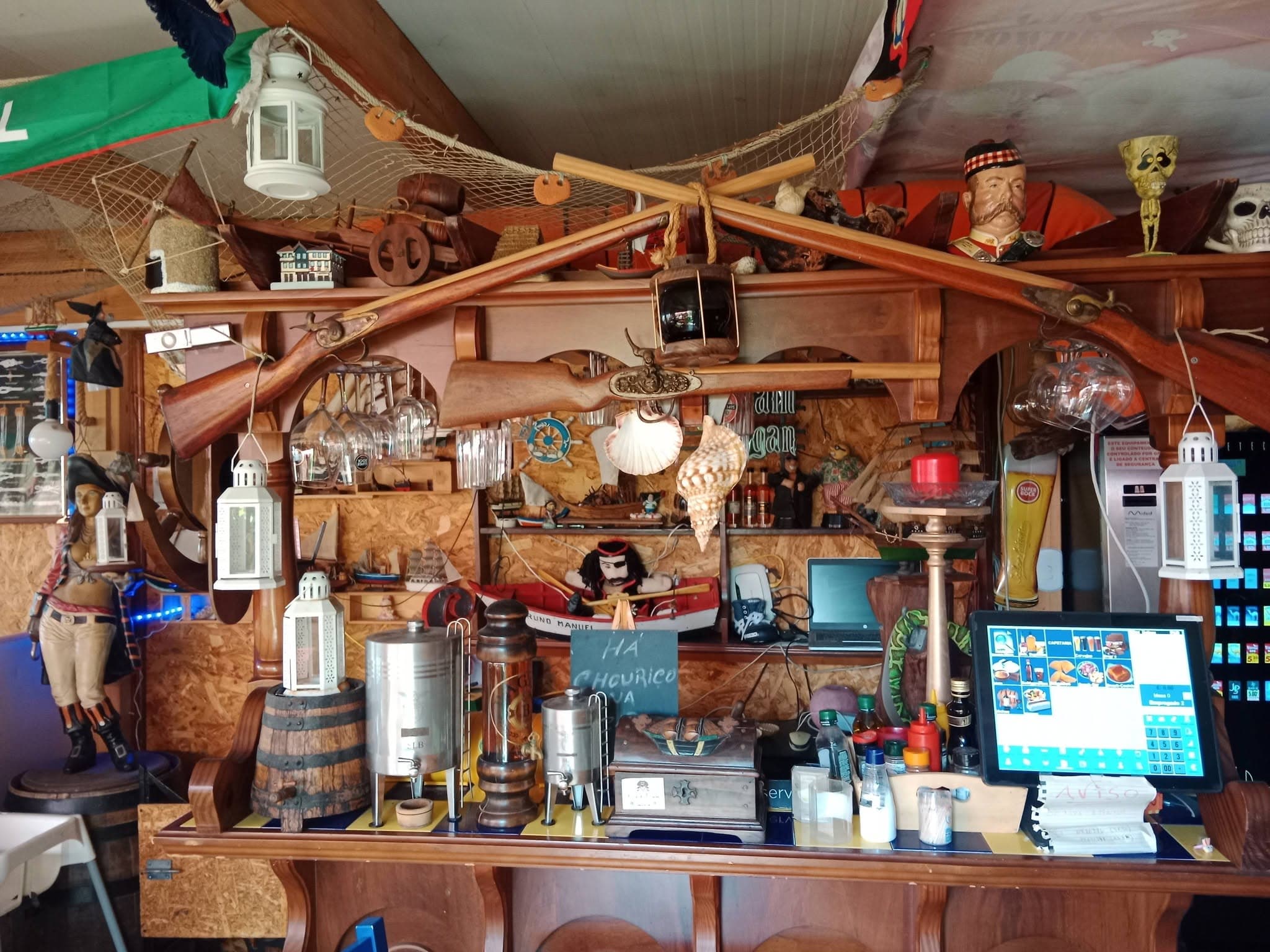 Duas espingardas antigas cruzadas com remos por cima do balcão, entre redes de pesca, lanternas brancas, conchas de vieira e uma fila de barcos de madeira.