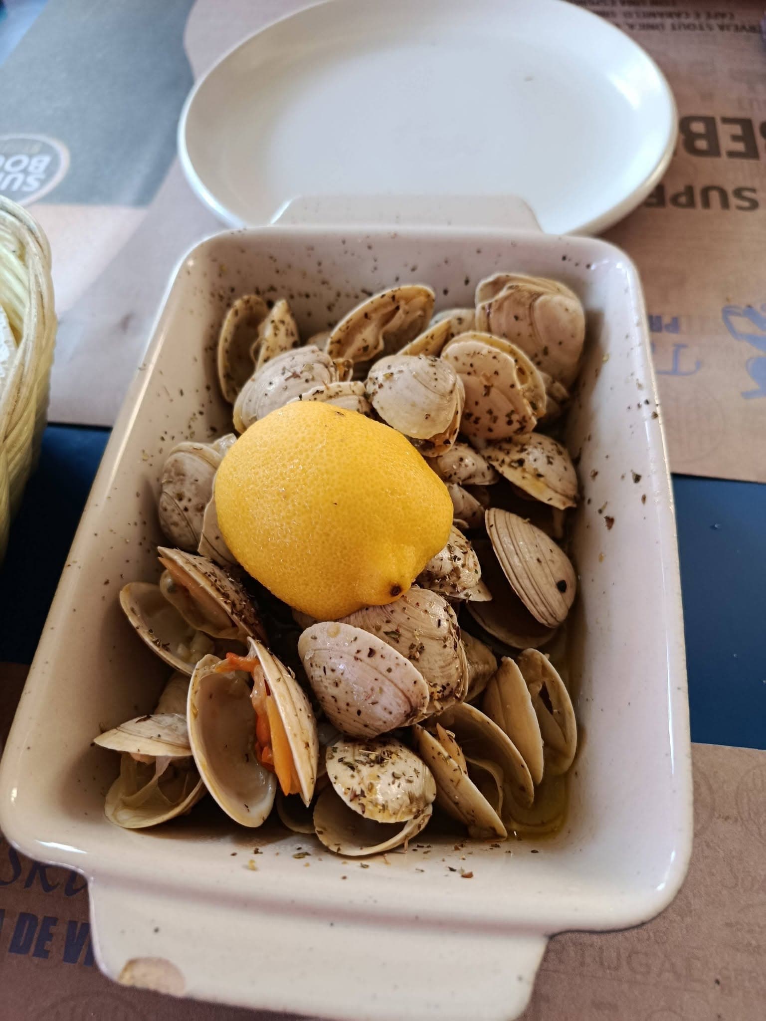 Uma travessa de amêijoas abertas em azeite e orégãos, com meio limão ao meio e um prato de pão ao lado.