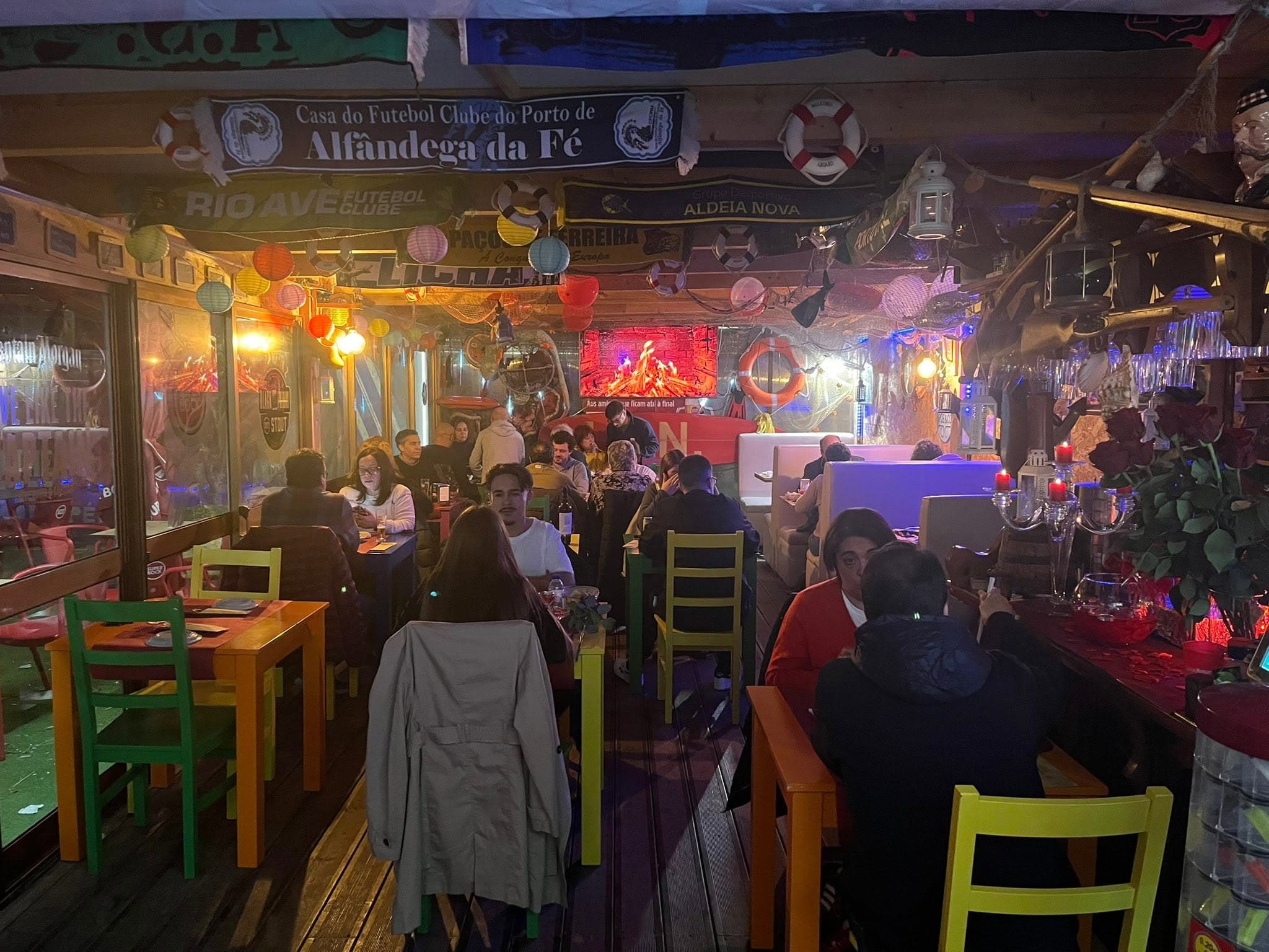 A sala cheia a uma noite de jantar, com cachecóis de clubes de futebol pendurados no tecto de madeira, lanternas de papel, bóias salva-vidas e mesas de cadeiras verdes, amarelas e laranja.